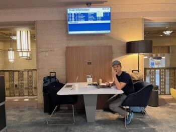 Jedd working at a table under train status screen in lounge